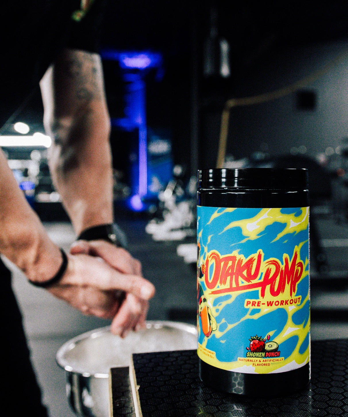 A tub of preworkout labeled Otaku Pump Shonen Punch with someone chalking their hands in the background in front of a gym setting